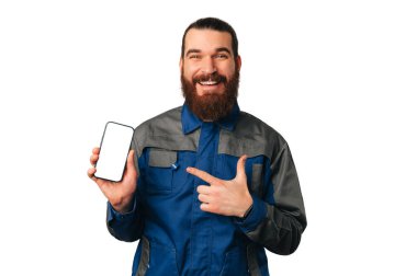Üniformalı gülümseyen tamirci, beyaz arkaplan üzerinde boş ekran olan bir telefona işaret ediyor..
