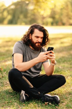 Uzun saçlı sakallı adam çimlerin üzerinde oturuyor ve güneşli bir günde parkta telefonunu kullanıyor..