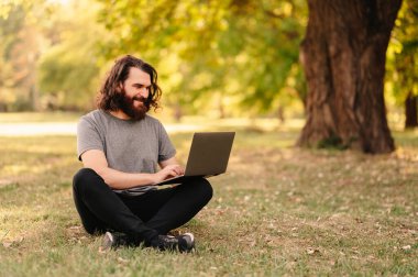 Uzun saçlı yakışıklı adam parkta çimlerin üzerinde otururken dizüstü bilgisayarını kullanıyor..