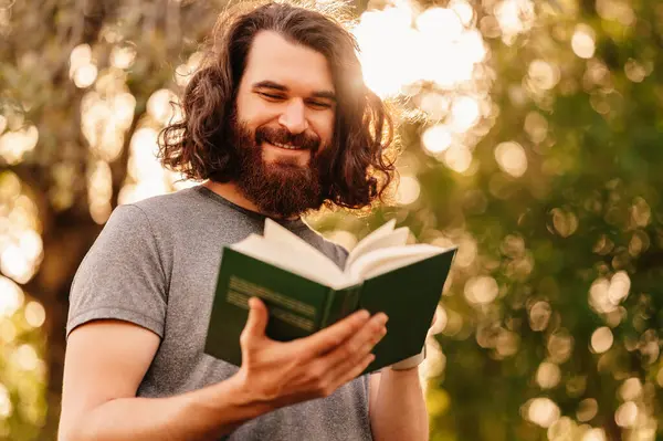 Uzun saçlı neşeli sakallı adam yazın parkta ilginç bir kitap okuyor..