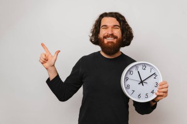 Neşeli sakallı adam elinde beyaz yuvarlak bir saat tutarken beyaz arka planı işaret ediyor..