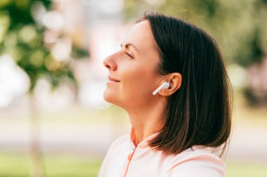 Açık havada gülümseyen orta yaşlı bir kadının güzel bir günün tadını çıkarırken kulaklık taktığı portreyi kapat..