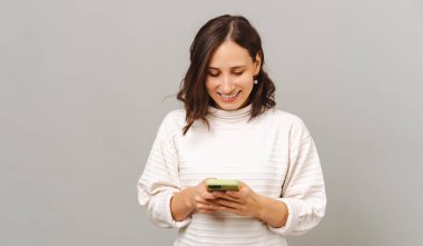 Telefonunu gri arka planda kullanan neşeli bir kadının stüdyo portresi.