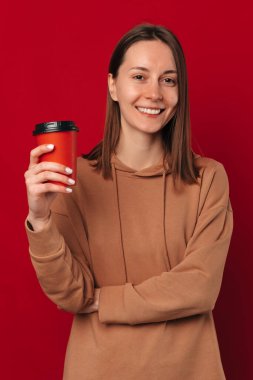 Kırmızı arka planda elinde bir fincan kahve tutan genç bir kadının dikey stüdyo fotoğrafı..