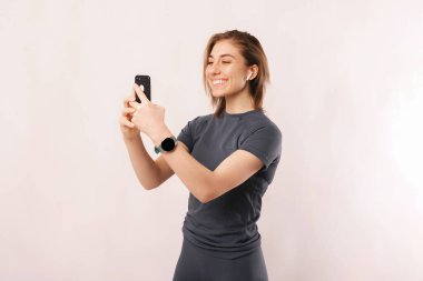 Spor kıyafetli, telefon ve kulaklıkla gülümseyen genç bir kadının stüdyo portresi. Selfie çeker ya da video görüşmesi yapar..