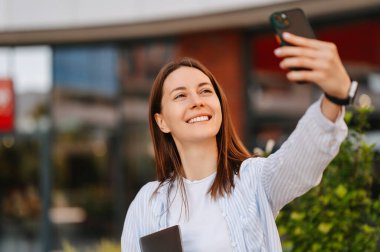Açık havada, akıllı telefonuyla selfie çeken neşeli genç bir kadının portresi..
