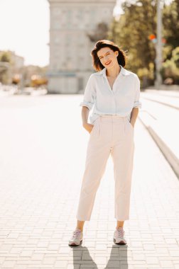 Güneşli bir günde, sokakta elleri ceplerinde duran genç bir kadının dikey dış görünüşü..