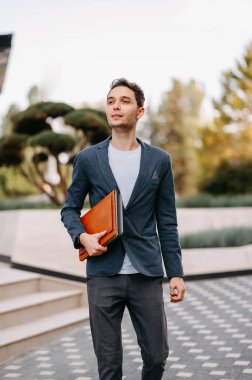 Ceket giyen genç bir adamın dikey portresi elinde dizüstü bilgisayarla dışarıda yürüyor..