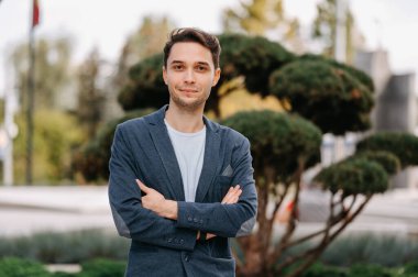 Kollarını kavuşturup kameraya bakan kendine güvenen genç bir adamın açık hava portresi..