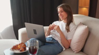 Genç ve neşeli bir kadının koltukta otururken dizüstü bilgisayarında video görüşmesi yaptığı video..