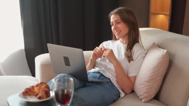 ソファーに座っている間にノートパソコンでビデオ通話をする若い陽気な女性のビデオ — ストック動画