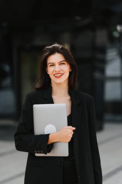 Dışarıda yürürken elinde dizüstü bilgisayarını tutan geniş gülümseyen bir kadının dikey portresi..