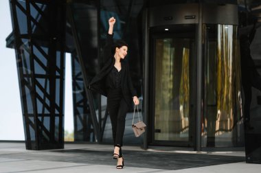 Takım elbiseli genç bir kadın camdan bir binadan çıkıyor sevinçten zıplıyor ve kazanan hareketi yapıyor..