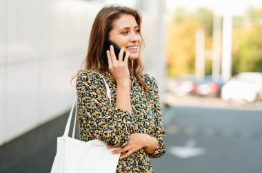 Elinde çantasıyla telefonla konuşan neşeli genç bir kadının açık hava portresi..