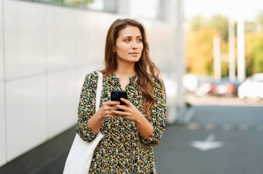 Açık havada yürürken telefonunu tutan güzel bir kadının portresi. Çiçekli bir elbise giyiyor ve bir çanta taşıyor..
