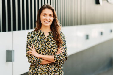 Açık havada, kollarını kavuşturmuş çiçekli elbiseli neşeli genç bir kadının fotoğrafını çek..