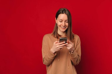 Kırmızı arka planda telefonunu kullanan genç bir kadının stüdyo portresi..