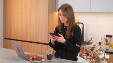 Genç bir kadın mutfakta dizüstü bilgisayarın önünde oturuyor ve akıllı telefondan mesaj yazıyor..