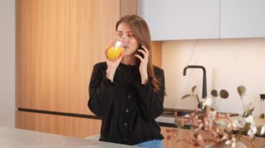 Mutfakta gülümseyen genç bir kadın akıllı telefondan konuşuyor ve bir bardak portakal suyu içiyor..