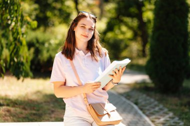 Genç neşeli kız öğrenci kitaplarıyla parkta ya da kampüste yürüyor..