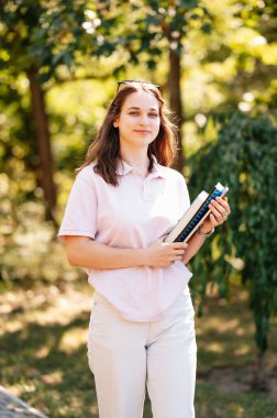 Neşeli kız öğrenci kampüste yürürken kitaplarını taşıyor. Kameraya gülümsüyor..
