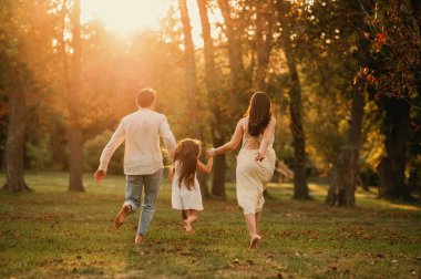 Üç kişilik genç bir aile el ele tutuşuyor ve parkta çıplak ayakla koşuyorlar. Arka plan görüntüsü.