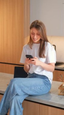 Mutfakta oturan, akıllı telefonunu kullanan ve gülümseyen bir kadının dikey videosu..