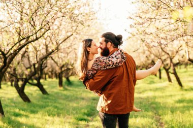 Genç yakışıklı adam, güneşli bir günde sevgilisini bir badem bahçesinde kollarıyla tutuyor..