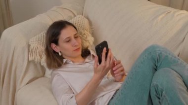 Bir kadın rahat bir evdeki koltukta rahat bir şekilde dinleniyor, akıllı telefonunu kullanıyor, modern yaşam tarzını somutlaştırıyor.