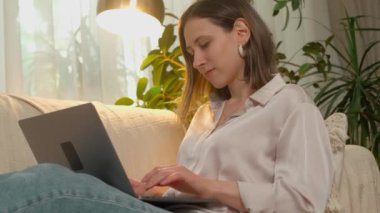 Hafif bluzlu genç bir kadın rahat bir ev ortamında rahat bir kanepede otururken dizüstü bilgisayarında daktilo kullanıyor..