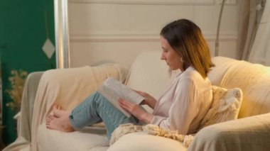 Genç bir kadın, rahat bir oturma odasında, rahat bir kanepede, bir kitaba sarılmış, huzur içinde dinleniyor.