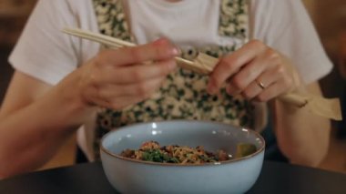 Restoranda yemek çubukları kullanarak, beyaz gömlek ve çiçekli önlük giyerek Asya mutfağından keyif alan bir adam..