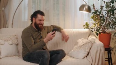 Bir adam rahat bir şekilde evdeki bir koltukta oturuyor, gülümsüyor ve arka planda oturma odası olan bir akıllı telefon kullanıyor.