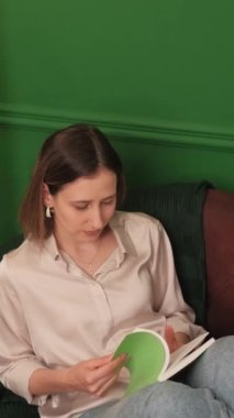 Yeşil duvarları ve rahat mobilyaları olan rahat bir odada, kanepede oturmuş yeşil bir kitap okuyan bir kadın..