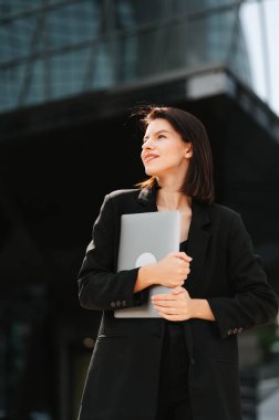 Kendine güvenen iş kadını elinde dizüstü bilgisayarla modern ofis binasının önünde dikiliyor.