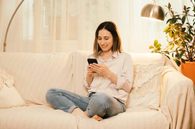 Genç bir kadın evinde kanepede dinleniyor, akıllı telefonuyla mesajlaşırken gülümsüyor, mutlu ve mutlu görünüyor.