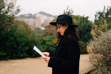 Açık havada gazete okuyan, siyah şapkalı ve paltolu bir kadının yan profili.