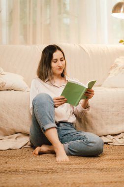 Bir kadın sıcak, rahat bir oturma odasında huzurlu bir atmosfer sergileyerek huzurlu bir şekilde kitap okumaktan zevk alır.