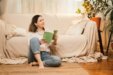 Bir kadın sakin bir şekilde oturma odasının zemininde oturuyor, bir kitaba dalmış, huzurlu bir anın tadını çıkarıyor.