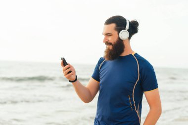 Mavi gömlekli genç bir adam kulaklıkla müzik dinliyor, gülümsüyor ve deniz kenarındaki akıllı telefonuna bakıyor..