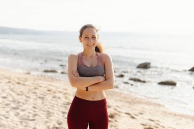 Genç bir kadın güneşli bir sahilde spor kıyafetleriyle kameraya poz verip gülümsüyor.