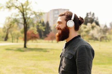 Sakallı bir adam, kulaklık takıyor, parkta geziniyor, kendini müziğe, doğaya ve güneşli bir güne kaptırıyor.