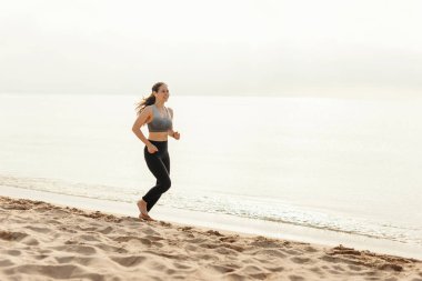 Bir kadın okyanusun kenarındaki kumlu bir sahilde koşu yaparken spor yapmayı ve açık havada sağlıklı bir yaşam sürmeyi teşvik ediyor.