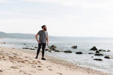 Kumlu sahilde, bir adam kayalık sahilde duruyor, doğal güzelliklerle çevrili sakin okyanus dalgalarına bakıyor.