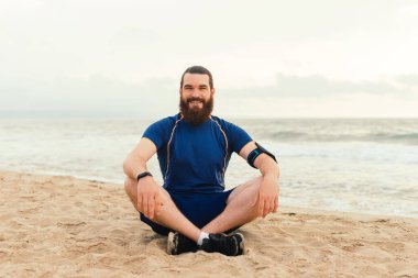 A bearded man in athletic clothing is seated on a sandy beach by the ocean, experiencing a tranquil moment clipart