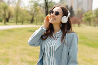 Bir kadın parkta gülümsüyor, kulaklıkla müzik dinliyor ve güneşli bir günde güneş gözlüğü takıyor.