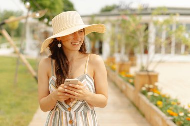 Güneşli bir açık havada akıllı telefonunu kullanırken şık bir yaz elbisesi ve şapkası içinde gülümseyen bir kadın..