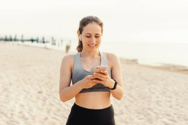 Spor kıyafetleri giyen bir kadın kumsalda akıllı telefonunu kullanırken görülüyor. Spor, yaşam tarzı ve açık hava teması için mükemmel.