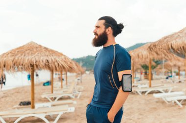 A man with a beard and bun is working out on the beach with a phone on his armband, enjoying the view clipart