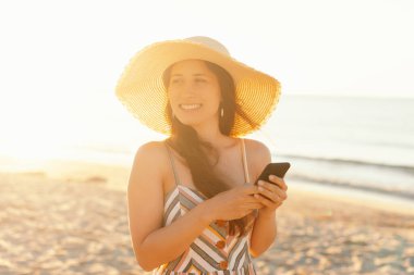 Geniş kenarlı şapkalı gülümseyen bir kadın gün batımında kumsalda telefonunu kullanarak güzel manzaranın tadını çıkarıyor.